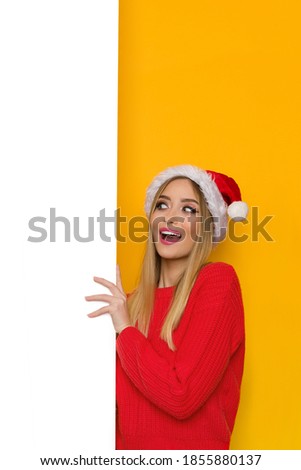Happy young woman in santa hat is holding white vertical banner, looking at it and talking. Waist up studio shot on yellow background.