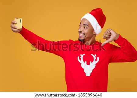 Funny young Santa african american man in Christmas hat doing selfie shot on mobile phone pointing thumb on himself isolated on yellow background studio portrait. New Year celebration holiday concept
