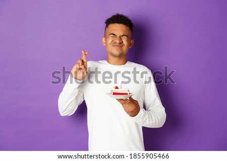Image of excited african-american man celebrating birthday, making wish, holding b-day cake and crossing fingers, standing over purple background