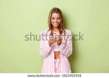 Image of beautiful blond woman in pink shirt, looking at mobile phone and smiling, standing over green background