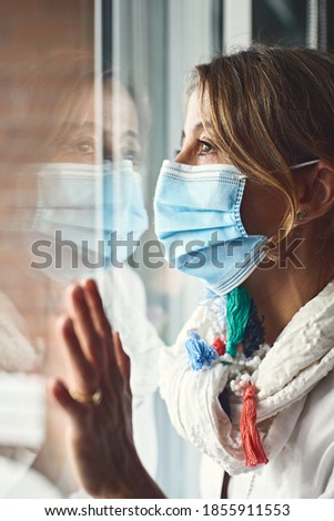 Middle-aged woman confined to home by covid-19, looks behind the glass. Confinement and covid-19. Protection mask against the virus.