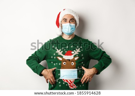 Concept of covid-19 and Christmas holidays. Funny guy put on face mask on his sweater deer, standing over white background