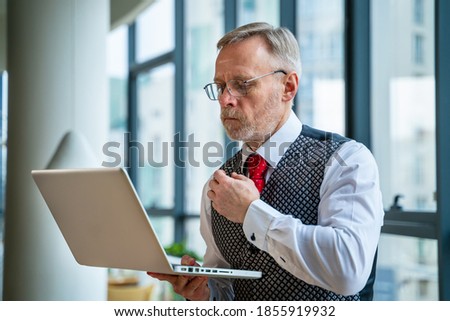 Mature businessman looking at laptop reading and printing improvements sitting near the window in his office. Panoramic city view background. Business concept.