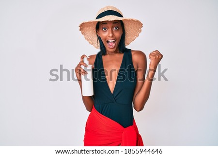 Young african american woman wearing swimsuit and holding sunscreen lotion screaming proud, celebrating victory and success very excited with raised arms 