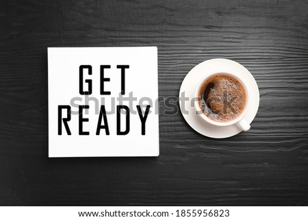Card with text Get Ready and cup of coffee on dark wooden table, flat lay
