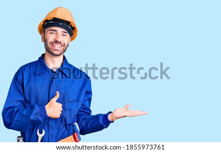 Young hispanic man wearing worker uniform showing palm hand and doing ok gesture with thumbs up, smiling happy and cheerful 