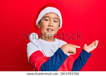 Little boy hispanic kid wearing christmas hat amazed and smiling to the camera while presenting with hand and pointing with finger. 