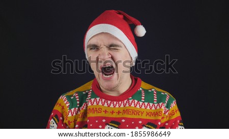 Young handsome guy shows emotion - yawn, he is wearing a red New Year's sweater with New Year's drawings, and a red New Year's hat