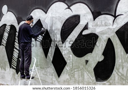 Graffiti artist in action, standing on ladders  and wearing protective face mask / respirator with filters. Abstract drawing with spray paint in a can on the wall. Street art culture concept.	