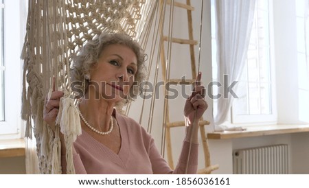 Calm pensioner woman wearing nice white pearl jewelry and pink jacket sits and swings in hammock by windows closeup slow motion