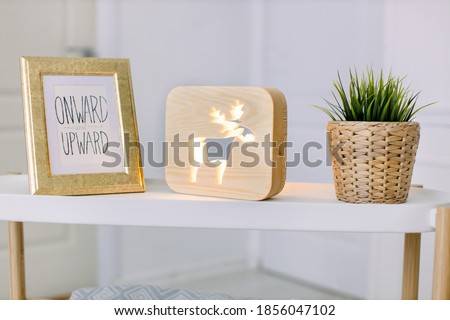 Design composition of modern home decor. Stylish table in light living room, with vintage photo frame, decorative wooden lamp with deer picture, and artificial plant in wicker flower pot