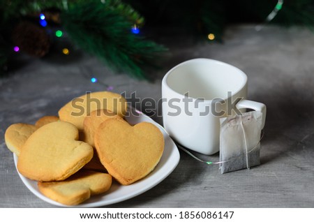 Homemade cookies in the shape of a heart in a white plate with a mug and a tea bag on the background of Christmas decorations.