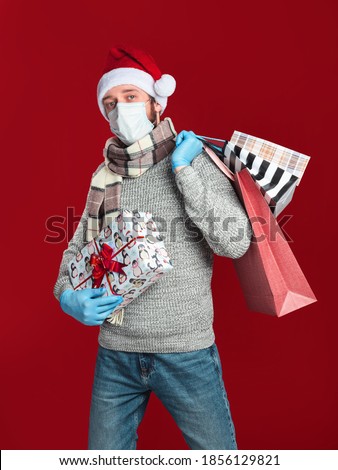 A young man in a medical mask and gloves, wearing a Santa hat and scarf holds a gift box and shopping bags on a red background