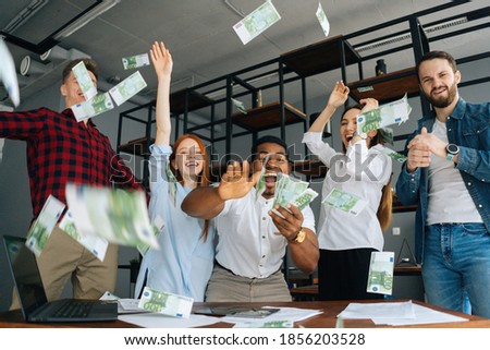 Low-angle shot of cheerful multi-ethnic employees celebrating victory and big profit at office workplace. Cheerful excited young business man and woman throwing away banknote. Rain of money