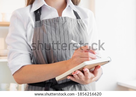 Culinary recipe. Festive bakery. Homemade food. Family traditional cuisine. Woman in cook apron writing in notebook light kitchen interior background.