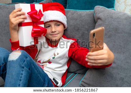 Handsome kid boy in Santa Claus hat looks at smartphone screen, shows a christmas present.Talk with family,closeup image.