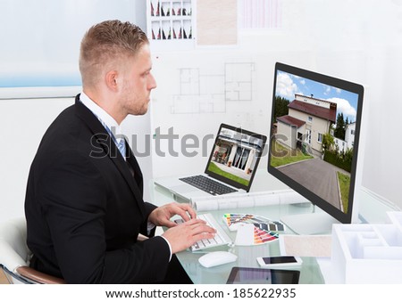 Businessman or estate agent checking a property portfolio online while sitting at his desk in the office looking at the exterior of a rural house visible on the desktop monitor