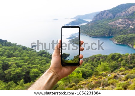 A hand with a smartphone taking pictures of the natural landscape, sea and mountains on a journey. Phone, internet and mobile network concept. Impressions and Adventures.