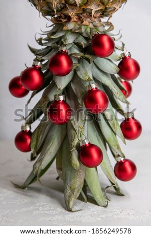 Original Christmas tree concept. Red shiny glass baubles hangs on the leaves of ripe pineapple standing upside down. Gray concrete festive background. Selective focus.