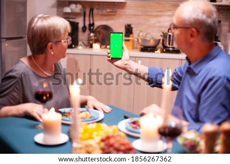 Old retired senior couple holding mockup phone at dinner. Aged people looking at mockup template chroma key isolated smart phone display using techology internet sitting at the table in kitchen.
