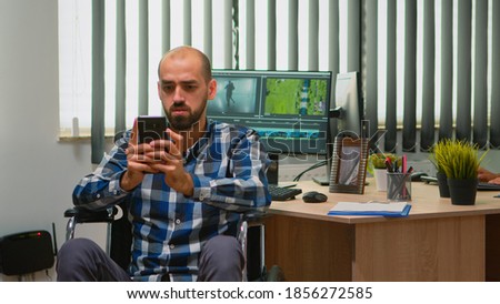 Freelancer photo designer with disability in wheelchair texting on smartphone taking break from editing video project creating content in modern company office. Blogger working from photo studio.