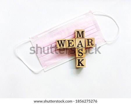 Top view photo of the square wooden blocks with crossword WEAR MASK on a pink disposable mask, isolated on white background.