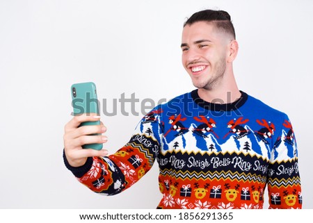Isolated shot of pleased cheerful Young handsome Caucasian man wearing Christmas sweater against white wall, makes selfie with mobile phone. People, technology and leisure concept