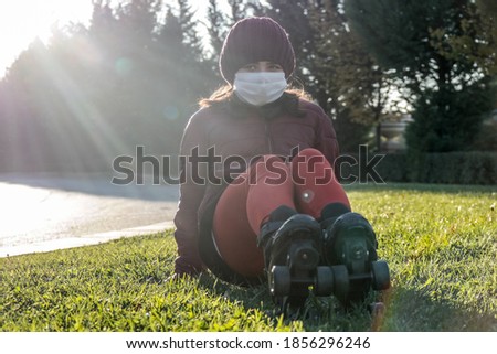 Primary school girl with red clothes, burgundy winter coat and knit hat with roller skates having a break on the grass with her face mask on for protection against Covid-19 outbreak
