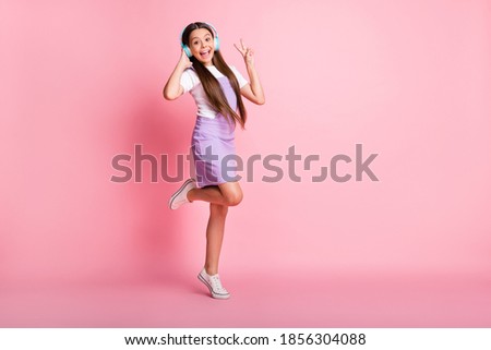 Full length photo of excited kid girl listen wireless headset music make v-sign isolated over pastel color background