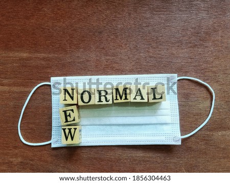 Top view photo of the square wood blocks crossword with the words NEW NORMAL on a disposable mask, placed on wooden ground.