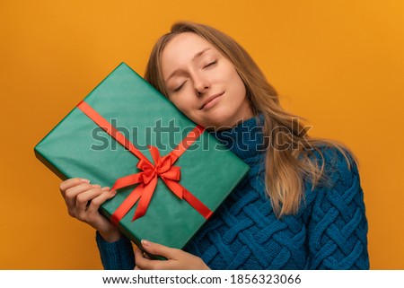 Image of charming young woman in warm knitted sweater smiling and holding gift with red ribbon. Studio shot, yellow background. New Year, Women's Day, Birthday, Holiday concept