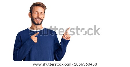 Handsome blond man with beard wearing casual sweater smiling and looking at the camera pointing with two hands and fingers to the side. 