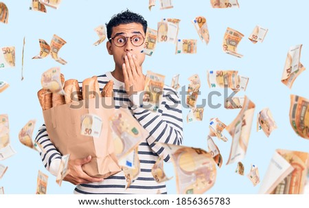 Hispanic handsome young man holding paper bag with bread covering mouth with hand, shocked and afraid for mistake. surprised expression