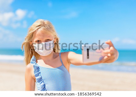 Funny girl taking selfie photo by smartphone on tropical sea beach. New rules to wear cloth face covering mask at public places due coronavirus COVID 19. Family holidays with children, summer travel.