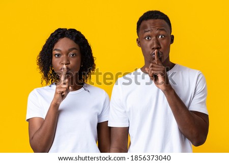Black couple make silence sign, keep forefingers on lips. Millennial african-american man and woman holding fingers on lips and looking at camera mysteriously over yellow studio background