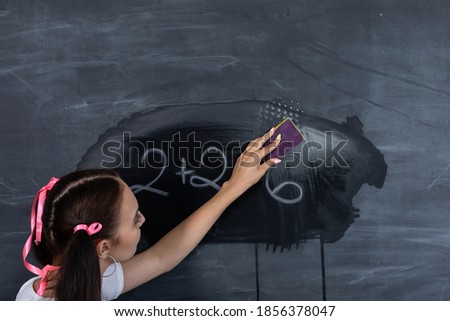 Before the next lesson, the girl wipes the chalkboard with a wet sponge. Her hair is pinned with a pink ribbon in two ponytails.