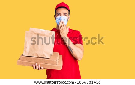 Young handsome hispanic man delivering food wearing covid-19 safety mask covering mouth with hand, shocked and afraid for mistake. surprised expression 