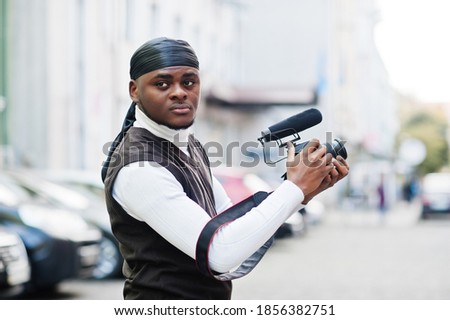 Young professional african american videographer holding professional camera with pro equipment. Afro cameraman wearing black duraq making a videos.