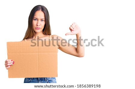 Beautiful brunette young woman holding cardboard banner with blank space with angry face, negative sign showing dislike with thumbs down, rejection concept 