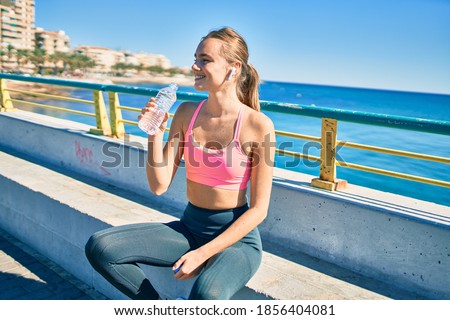 Young blonde sportswoman drinking bottle of water sitting on the bench at the promenade.