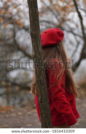 Little sad long haired girl in red hat and coat turned away and leaned to tree. Bokeh effect.