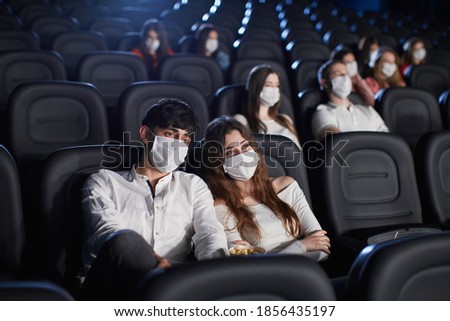 Selective focus of man and woman enjoying film during world pandemic, looking at screen. Young caucasian audience watching movie in cinema, wearing white face masks. Social distancing concept. Royalty-Free Stock Photo #1856435197