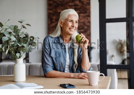 Happy senior woman eating healthy green apple in kitchen Royalty-Free Stock Photo #1856473930