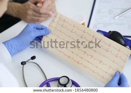 Doctor's hands hold result of the cardiogram next to patient sitting. Examination of the cardiovascular system concept. Royalty-Free Stock Photo #1856480800