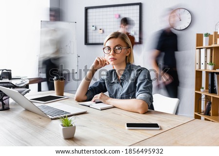 blonde businesswoman in eyeglasses sitting at workplace with gadgets in open space office, motion blur Royalty-Free Stock Photo #1856495932