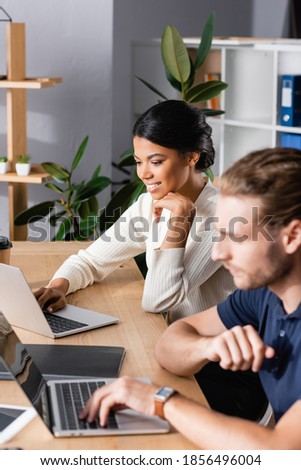 Smiling african american woman looking at laptop while sitting at workplace with blurred man on foreground