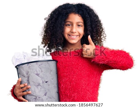 African american child with curly hair holding paper bin full of crumpled papers smiling happy and positive, thumb up doing excellent and approval sign 