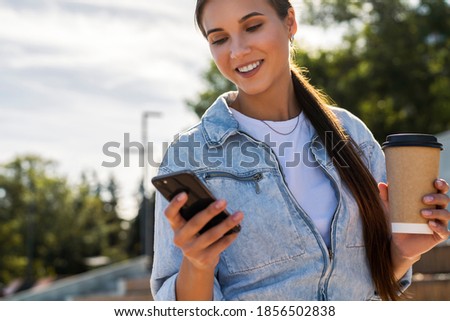 Attractive young girl in denim clothes writes messages using smartphone. Beautiful brunette woman is texting with friends on social networks while on street in public park. Woman communicates