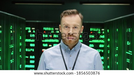Portrait of handsome Caucasian young man looking straight to camera in big data storage room with many processors and smiling. Male system administrator standing among servers in datacenter.