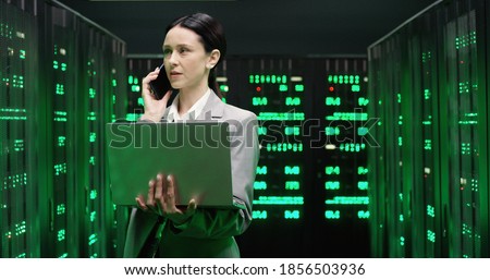 Caucasian beautiful woman walking among servers with laptop computer and talking on mobile phone. Pretty female analytic in data storage working on digital security and speaking on cellphone.